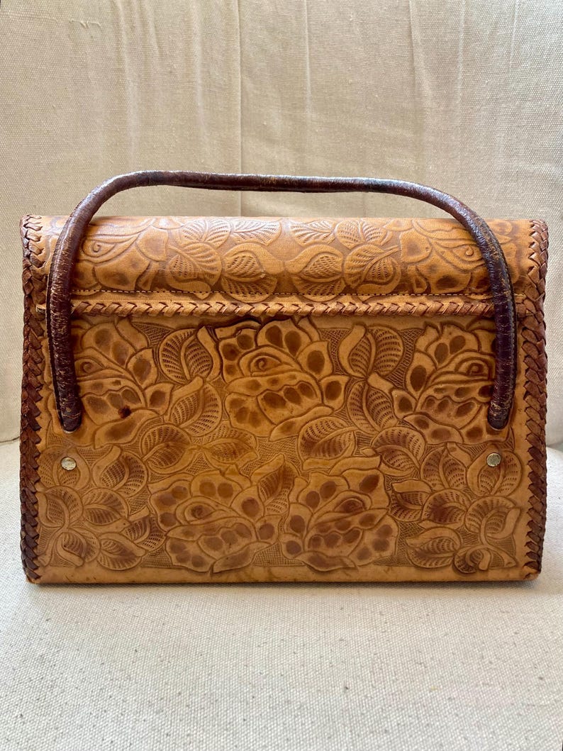 Pode incluir: Bolsa de m&atilde;o de couro castanho com um design floral em relevo. A bolsa apresenta uma al&ccedil;a e acabamentos em castanho escuro, com formato retangular e uma borda tran&ccedil;ada. O padr&atilde;o floral cobre a frente e as costas da bolsa.