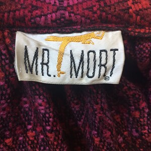 May include: A white label with black text that reads "MR. MORT" and a gold embroidered image of a stylized bird. The label is on a red and black patterned fabric.