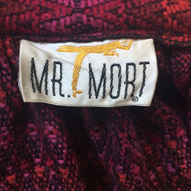 May include: A white label with black text that reads "MR. MORT" and a gold embroidered image of a stylized bird. The label is on a red and black patterned fabric.