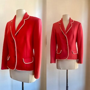 May include: A vibrant red blazer featuring a crisp white piped trim along the lapels, front opening, and two flap pockets. The jacket is structured with long sleeves and a button-front closure, displayed on a mannequin to show its tailored fit.