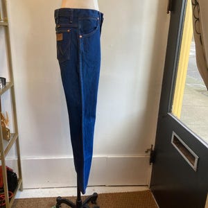 May include: Dark blue denim jeans with a classic five-pocket design. The jeans are displayed on a mannequin, showcasing the straight leg cut and the brown leather patch on the back pocket. The jeans have copper-colored rivets and stitching.