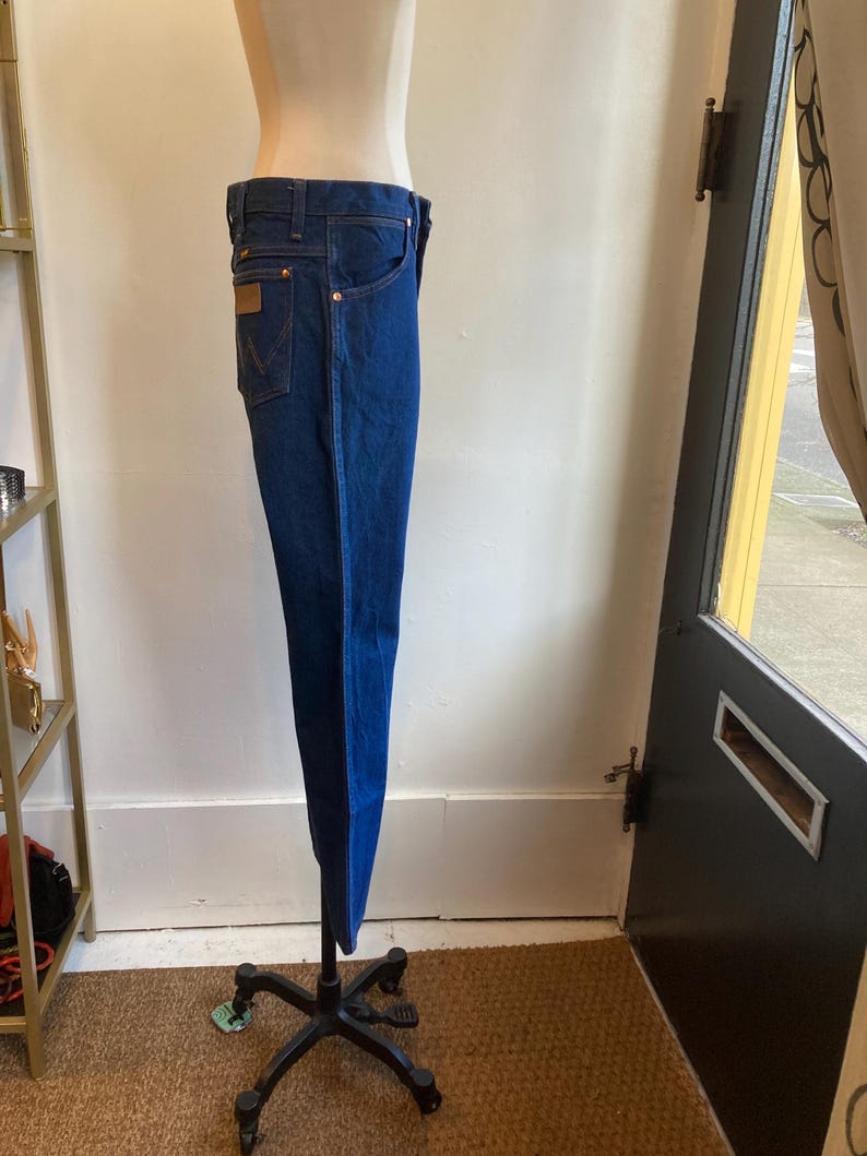 May include: Dark blue denim jeans with a classic five-pocket design. The jeans are displayed on a mannequin, showcasing the straight leg cut and the brown leather patch on the back pocket. The jeans have copper-colored rivets and stitching.