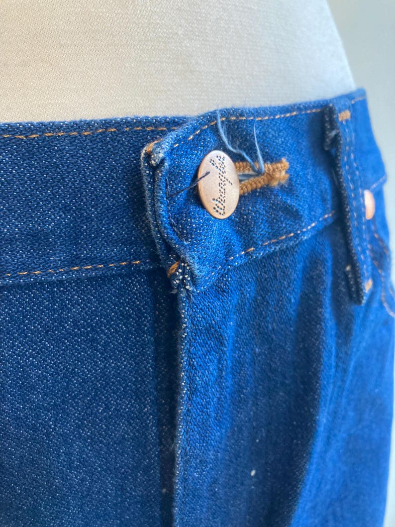 May include: Close-up of the waistband of blue denim jeans. The jeans have a button closure with the word "Wrangler" engraved on the button. The stitching is a contrasting tan color.
