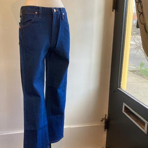 May include: Dark blue denim jeans with a classic five-pocket design. The jeans are displayed on a mannequin, showcasing their straight leg fit and copper-colored rivets. The jeans have a button and zipper closure.