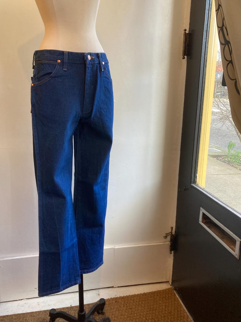 May include: Dark blue denim jeans with a classic five-pocket design. The jeans are displayed on a mannequin, showcasing their straight leg fit and copper-colored rivets. The jeans have a button and zipper closure.