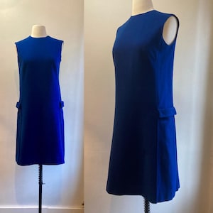 May include: A vibrant royal blue sleeveless dress with a round neckline and two small pockets. The dress is displayed on a mannequin, highlighting its simple, elegant design. The dress has a straight silhouette.
