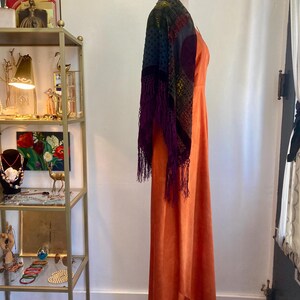 May include: A long, burnt orange maxi dress with a V-neckline. The dress is made of a silky fabric and has a flowing silhouette. A colorful shawl with fringe is draped over the dress.