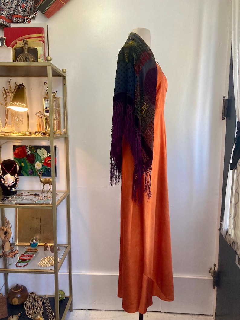 May include: A long, burnt orange maxi dress with a V-neckline. The dress is made of a silky fabric and has a flowing silhouette. A colorful shawl with fringe is draped over the dress.