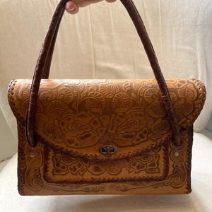 Pode incluir: Uma mala de m&atilde;o de couro castanho com um design floral em relevo. A mala tem uma forma retangular com fecho de aba e duas al&ccedil;as castanhas. A mala &eacute; segurada por uma pessoa.
