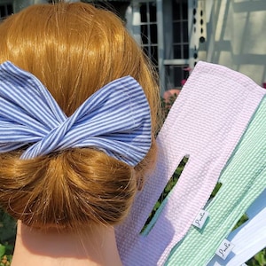Könnte beinhalten: Eine blaue und weiße gestreifte Stoffschleife ist im Haar einer Frau gebunden. Die Schleife besteht aus einem weichen, strukturierten Stoff. Die Frau hält drei Stoffhaarbänder in der Hand. Die Haarbänder sind in rosa, grün und weiß. Jedes Haarbänder hat ein weißes Etikett mit dem Wort "Pale" darauf.