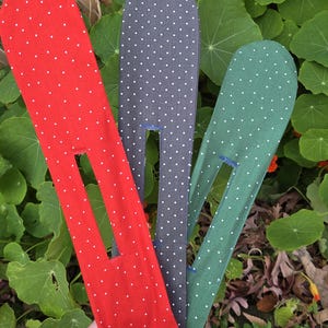 Op de afbeelding: Drie stoffen haaraccessoires in rood, grijs en groen, elk met witte stippen. De accessoires hebben een rechthoekige opening en worden in een hand gehouden tegen een achtergrond van groene bladeren en oranje bloemen.