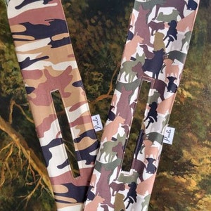 May include: Two fabric headbands with a camouflage pattern. One headband features a brown, tan and black camouflage pattern. The other headband features a brown, tan and black camouflage pattern with a silhouette of dogs printed on it. Both headbands have the word "Pawle" printed on them.
