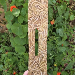 May include: A wooden decorative item with a cutout, featuring a repeating pattern of swirling lines and dots in shades of brown and beige. The item is held in front of a backdrop of green foliage and orange flowers.