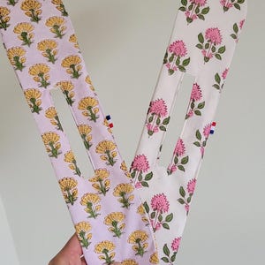 Pode incluir: Dois scrunchies de tecido com padrões florais. Um scrunchie é rosa com flores amarelas e o outro é branco com flores rosas.