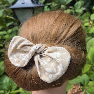 Könnte beinhalten: Ein Haaraccessoire bestehend aus einer beigen Stoffschleife mit weißem Blumenmuster. Die Schleife ist in der Mitte geknotet und liegt horizontal auf einem Dutt aus rotbraunem Haar, wobei sie das Haar durch ihre Stoffkonstruktion zusammenhält.