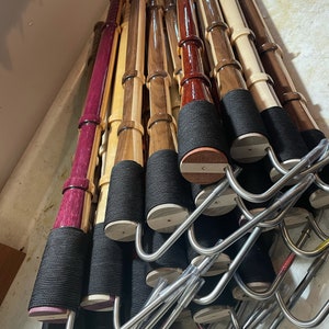 Può includere: Una collezione di bastoni da passeggio in legno di vari colori, tra cui viola, marrone chiaro e marrone scuro. Ogni bastone ha un'impugnatura intrecciata nera e una base in legno. I bastoni sono esposti su ganci di metallo.