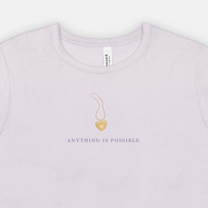 May include: A light purple t-shirt with a gold pendant necklace design and the phrase "ANYTHING IS POSSIBLE" printed below the necklace. The t-shirt is made of a soft material.