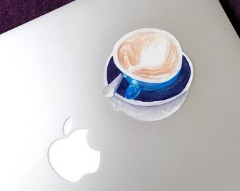 May include: A sticker of a latte with a heart-shaped design on top, in a blue cup and saucer. The sticker is placed on a silver laptop, with a purple background.