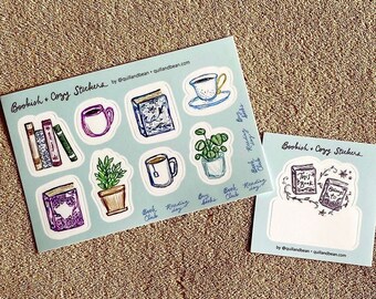 May include: Two sticker sheets with a light blue background and the text "Bookish + Cozy Stickers." One sheet features illustrations of books, mugs, and plants. The other sheet has illustrations of books and the text "This Book" and "Bring to Life."