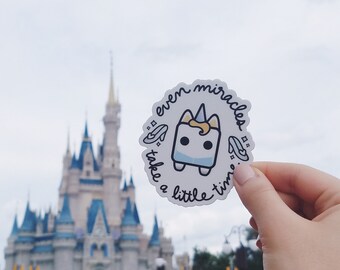 May include: A white sticker with a cartoon unicorn and the text "even miracles take a little time". The sticker is being held in front of Cinderella's Castle at Walt Disney World.