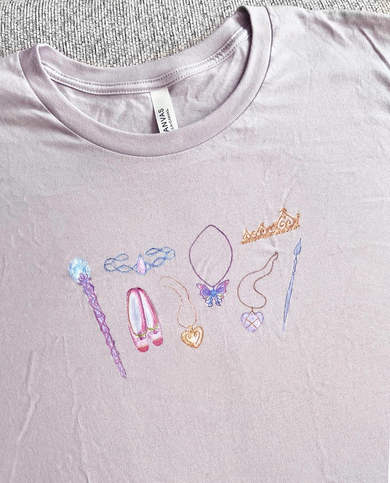 May include: A light purple t-shirt with a design of various princess-themed items, including a crown, wand, shoes, necklace, and butterfly. The design is embroidered on the shirt.