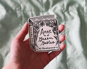 May include: A black and white sticker featuring the book "Anne of Green Gables" with floral accents. The title and author's name are visible. The sticker is held in a hand against a light green background.
