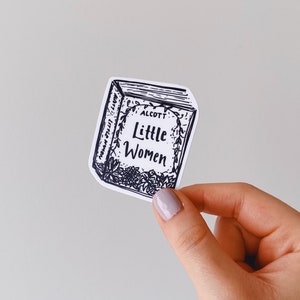 Puede incluir: Una pegatina en blanco y negro de una portada de libro con el título "Little Women" de Louisa May Alcott. La portada del libro está rodeada de un diseño floral.