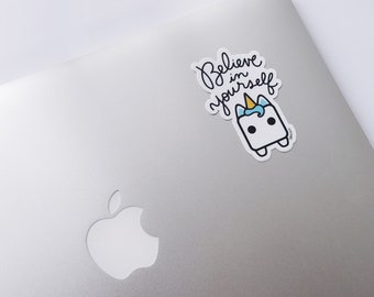 May include: A white sticker with the words "Believe in yourself" in black script. The sticker features a cartoon unicorn with a blue mane and a yellow horn. The sticker is on a silver laptop.