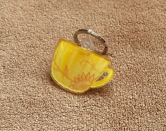 May include: A yellow teacup keychain with a floral design and a sunflower. The keychain is made of clear acrylic and has a silver metal ring.