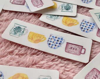 May include: White rectangular stickers with watercolor illustrations of mugs in various styles and colors. The mugs feature designs like floral patterns, polka dots, and mountain scenes. The stickers are scattered on a pink, fuzzy surface.