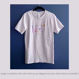 Puede incluir: Camiseta lila claro con cuello redondo, con una colorida ilustraci&oacute;n de accesorios. El dise&ntilde;o incluye una varita, zapatos, un collar y una corona. El texto "Live your own fairy tale" est&aacute; impreso debajo.