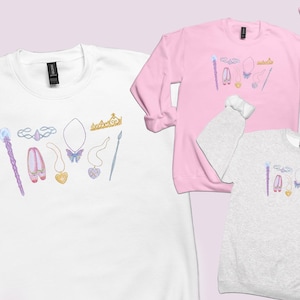 Puede incluir: Tres sudaderas de cuello redondo en blanco, rosa claro y gris jaspeado, cada una con un estampado gráfico central de una varita, tiara, zapatillas de ballet y varios colgantes en forma de corazón y mariposa en tonos pastel y dorados.