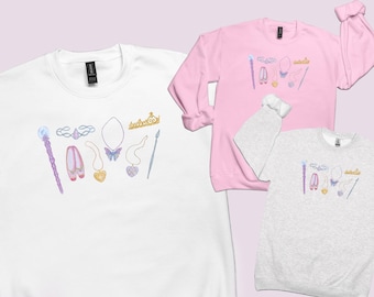 May include: Three crewneck sweatshirts in white, light pink, and heather gray, each featuring a central graphic print of a wand, tiara, ballet slippers, and various heart and butterfly-shaped pendants in soft pastel colors and gold tones.