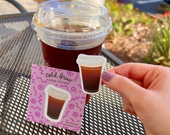 May include: A pink and purple sticker with a cartoon illustration of a cup of cold brew coffee. The sticker says "cold brew coffee sticker" and includes the social media handle @quilandbean and website quilandbean.com.