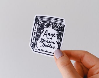 May include: A black and white sticker of the book cover for "Anne of Green Gables" by L.M. Montgomery. The cover features a stylized illustration of flowers and the title of the book.