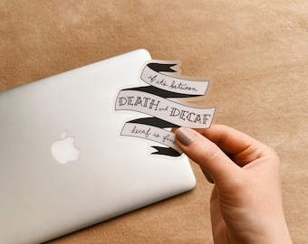 May include: A clear sticker with black text that reads "if it's between DEATH and DECAF, decaf is fine". The sticker is shaped like a banner and is being held by a hand over a silver laptop.