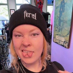May include: A person wearing a black beanie with the word "feral" embroidered on it. They are wearing a black t-shirt with a white graphic that says "Got girls read dark fantasy".