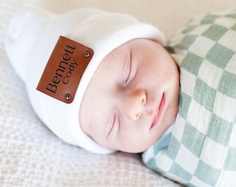 May include: A newborn baby wearing a white beanie with a brown leather tag that reads "Bennett Cody." The baby is wrapped in a green and white checkered blanket. The baby is sleeping peacefully on a white textured surface.