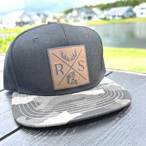 May include: A black baseball cap with a camouflage print brim. The front features a square brown leather patch with a black engraved design showing the letters R and S, a pair of deer antlers, and a fish illustration separated by an X.