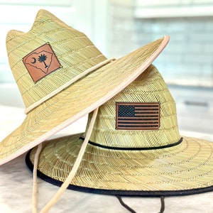 May include: Two straw sun hats with leather patches. The top hat has a brown patch with a white outline of the state of South Carolina and the words "South Carolina". The bottom hat has a brown patch with a white outline of the American flag.