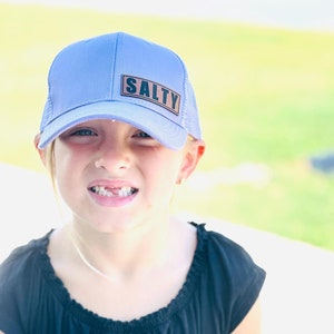 May include: A light blue baseball cap featuring a rectangular brown patch on the front panel with the word SALTY printed in bold black capital letters. The cap has a structured crown and a curved brim, designed with a mesh back for ventilation.