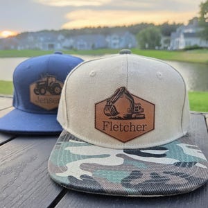 May include: Two baseball caps with leather patches. The front cap is beige with a camouflage brim and a patch featuring an excavator and the name Fletcher. The back cap is navy blue with a patch featuring a tractor and the name Jenser.