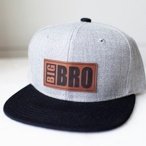 May include: A gray flat-brimmed baseball cap with a black brim. The cap features a brown leather patch with the words "BIG BRO" in black block letters. The cap is a stylish accessory.