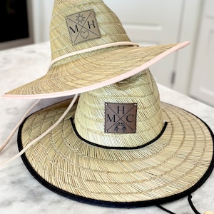 May include: Two straw hats with wide brims. The top hat has a pink trim and a leather patch with the letters "M" and "H" and a heart and sun symbol. The bottom hat has a black trim and a leather patch with the letters "H", "M", and "C" and a crab symbol.