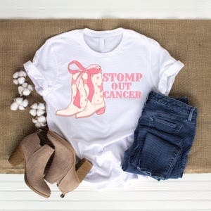 May include: White t-shirt with a pink and cream cowboy boot graphic and the text "STOMP OUT CANCER." The shirt is paired with blue jeans and tan ankle boots. The shirt is laying on a brown surface.