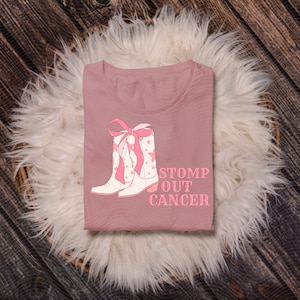 May include: A dusty rose t-shirt featuring a graphic of white cowboy boots with pink accents and a pink bow. The text "STOMP OUT CANCER" is printed in pink below the boots. The shirt is displayed on a white fluffy surface.