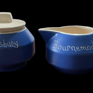 May include: A blue ceramic sugar bowl with a lid and a creamer. The sugar bowl has the word "Shaftesbury" in white script, and the creamer has "Bournemouth". Both pieces have a white rim and handle.
