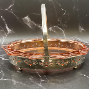 Peut inclure: Un panier ancien en verre rose et métal argenté avec une poignée. Le panier présente un design géométrique avec un bord et une base décoratifs. La poignée est arquée et fixée au bord. Le panier est surélevé sur de petits pieds.