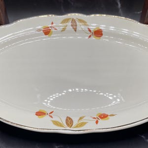 May include: An oval ceramic serving platter with a cream-colored base and a scalloped edge featuring a metallic gold trim. The rim is decorated with a repeating pattern of orange and yellow flowers accompanied by brown and tan leaves.
