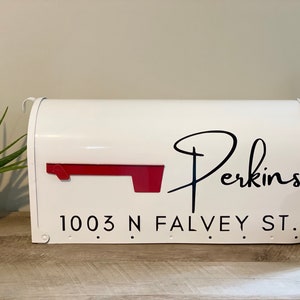 May include: White mailbox with a red flag and the name "Perkins" in black script. The address "1003 N Falvey St." is printed below. The mailbox is on a wooden surface, with plants and a jar to the sides.